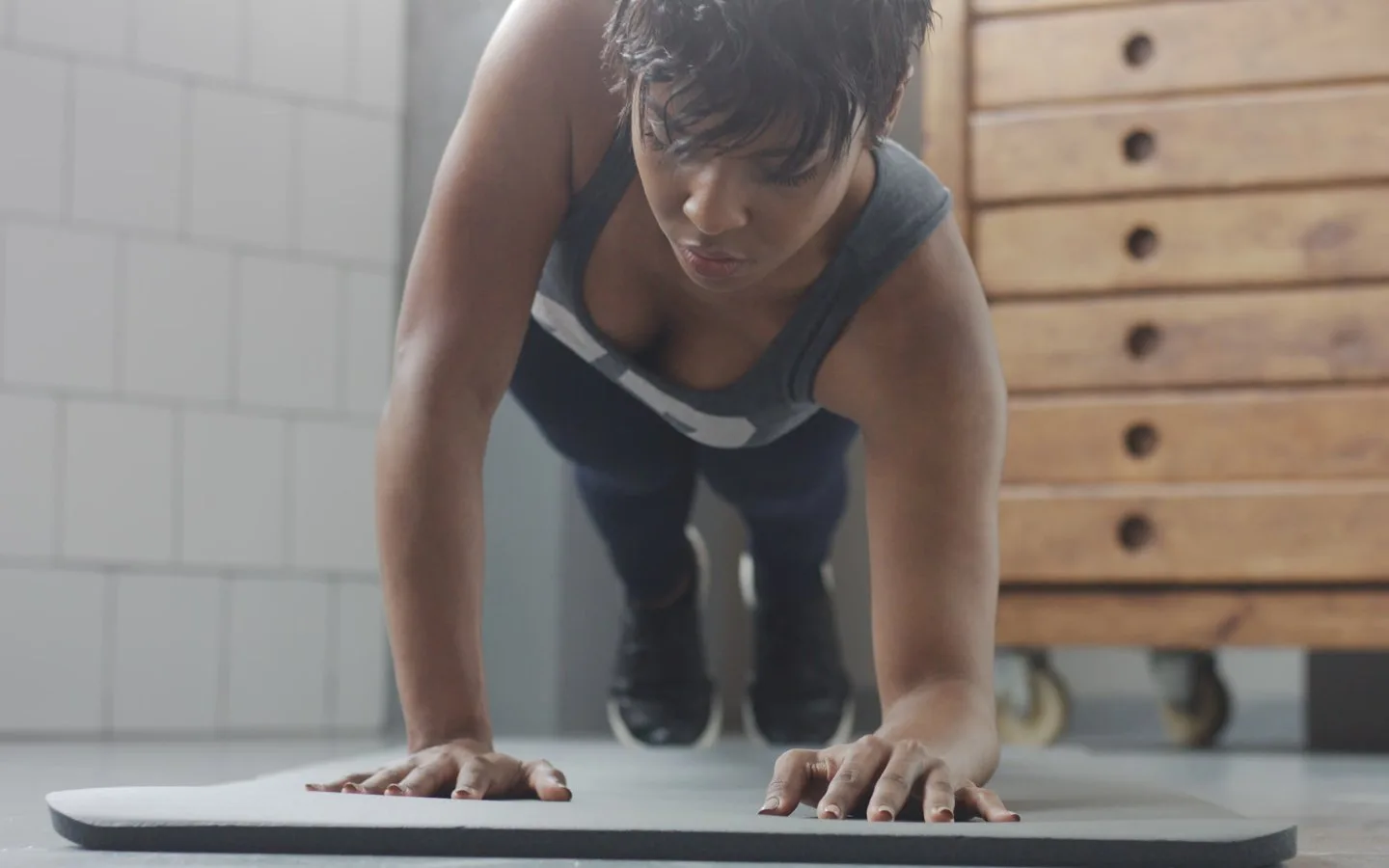 Una mujer con camiseta de tirantes hace una plancha sobre una esterilla en una sala de entrenamiento con pared de azulejos blancos y un mueble de cajones de madera.
