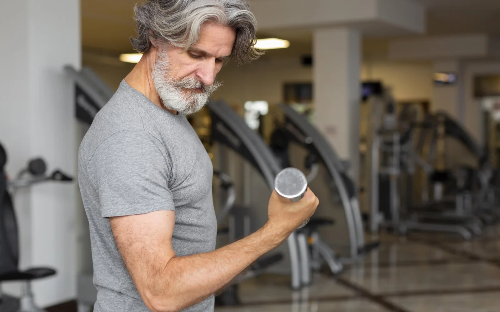 Hombre mayor de pelo y barba canosos haciendo un curl de bíceps con una mancuerna en un gimnasio, concentrado en el movimiento.
